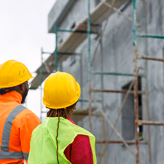 Two engineers wearing yellow hard hats and reflective vests observe scaffolding on a concrete building structure.