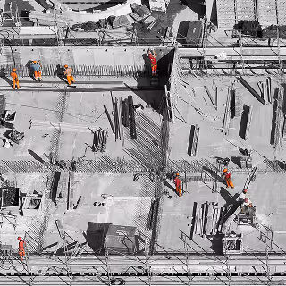 Top-down view of construction workers in orange safety gear navigating a complex worksite with exposed rebar and framework.
