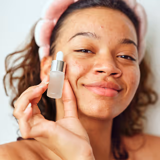 A young woman with natural skin and acne holds a small frosted glass serum bottle.
