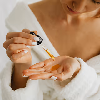 A person in a bathrobe drops golden face serum onto her palm with a dropper.