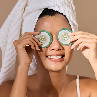 A smiling woman with a towel on her head holds green eye patches over her eyes.