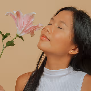 A serene woman gently smells a large, pink lily against a neutral background, symbolizing relaxation, tranquility, and the sensory pleasure associated with scent.