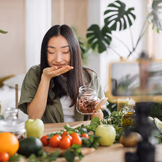 A smiling woman enjoys the aroma of ingredients from a jar, surrounded by fresh produce and herbs, in a bright, natural kitchen setting, suggesting a sense of freshness and wellbeing.
