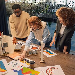 Three colleagues smiling and collaborating on a creative project around a table filled with colorful charts, graphics, and wind turbine models, reflecting teamwork and innovation.