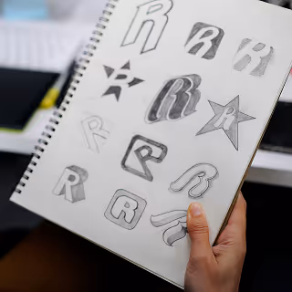 A hand holding a sketchbook filled with various hand-drawn letter "R" logo concepts, highlighting the process of iterative design and logo brainstorming.