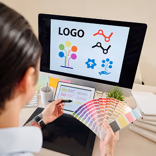 A graphic designer reviewing colorful digital logo concepts on a computer screen, holding a Pantone color guide, symbolizing the thoughtful selection of brand colors.