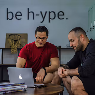 Two men comfortably engaging in a discussion around a laptop in a casual office space featuring modern decor and motivational branding, reflecting collaboration and strategic thinking.