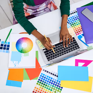 Overhead view of a designer working on a laptop surrounded by bright color swatches, design sketches, and a color wheel, illustrating creative color planning and brand identity development.