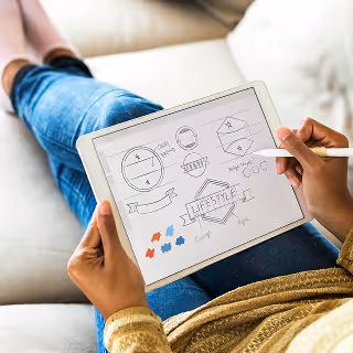 A person seated comfortably, using a stylus to sketch logos and design elements on a tablet screen, emphasizing creative digital branding processes.