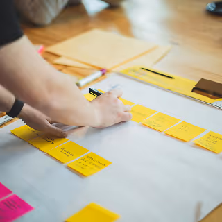 Close-up of a person's hand organizing bright yellow sticky notes on a large sheet of paper, suggesting structured planning and idea mapping for product strategy.
