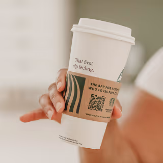 Person holding a coffee cup displaying branding and a QR code, representing practical brand implementation and consumer interaction.