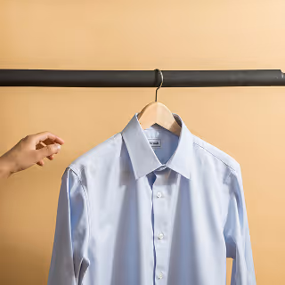 A neatly pressed, pale blue dress shirt hanging on a wooden hanger against a beige backdrop, with a hand reaching to adjust its position, emphasizing product presentation and attention to detail.