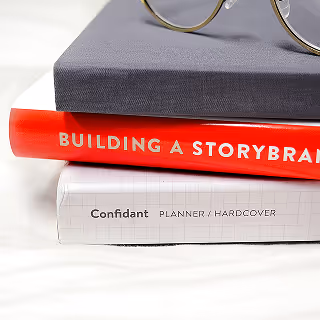 Close-up of a neatly stacked pile of books with visible titles including 'Building a Storybrand,' alongside a pair of eyeglasses on top.