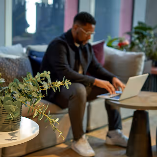 A man focused on working on his laptop in a comfortable, modern lounge area with natural elements and cozy furnishings.