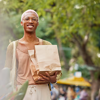A cheerful person with short pink hair and glasses, standing outdoors in a park setting, holding two brown paper shopping bags. They are dressed casually and smiling warmly.