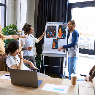 A group of young professionals gathered in an office, presenting colorful charts on a flip board. One person stands pointing at the charts while others sit and engage with the presentation.