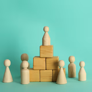 Wooden figurines arranged around a small pyramid made of wooden blocks. A single figure is placed at the top of the pyramid, symbolizing leadership or hierarchy, set against a mint green background.