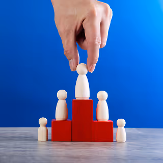 A hand placing a wooden figurine on top of red building blocks, surrounded by smaller wooden figures, symbolizing leadership, hierarchy, or promotion against a bright blue background.