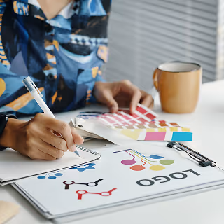 Person sketching in a notebook while reviewing colorful logo concepts and a Pantone color swatch, suggesting a branding or design brainstorming session.