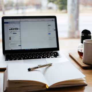 Open notebook with a pen resting on its pages in front of a laptop displaying a document editor, with a coffee mug and window view in the background, evoking a calm and focused work environment.