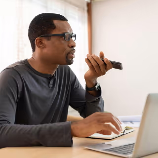 Man speaking into a smartphone using voice input while working at a laptop, with a notepad nearby, portraying multitasking in a remote or home office setting.