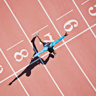 Aerial view of a sprinter in a light blue uniform crossing the finish line with arms raised in triumph on a pink track, symbolizing achievement and success.