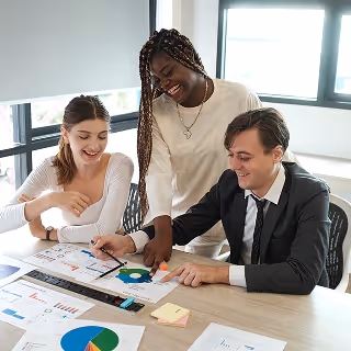 Three colleagues reviewing colorful data charts at a desk, smiling and collaborating in a bright office environment.