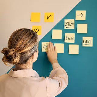 Woman placing sticky notes labeled with words like 'goal,' '2024,' and 'to do' on a teal and beige wall, illustrating goal-setting and strategic planning.