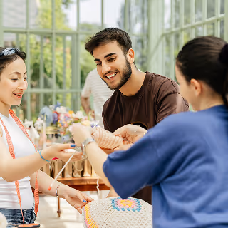 Three young adults smiling and collaborating on a crochet or handmade craft project in a bright greenhouse-style workspace, reflecting creative teamwork and community.