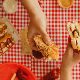 Hands holding sandwiches over a red and white checkered picnic table, surrounded by hot dogs, dips, and plates, suggesting casual dining or food-sharing moments.