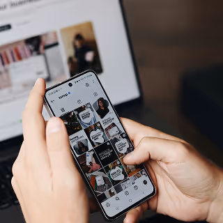 Person scrolling through a visually branded Instagram feed on a smartphone, with a laptop open in the background, indicating social media engagement.
