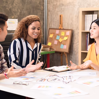 Three colleagues having a casual discussion at a desk covered in colorful charts and diagrams, with a corkboard and bookshelf in the background.