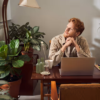 Young man seated at a cozy desk surrounded by plants, gazing thoughtfully away from his laptop, suggesting contemplation or creative thinking.