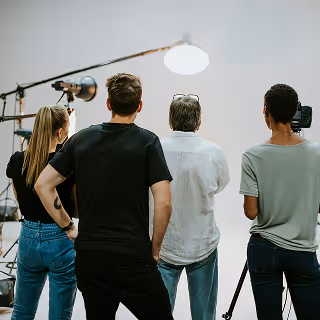 A small video production team, seen from behind, watching a shoot on set with studio lights and cameras arranged around a white backdrop.