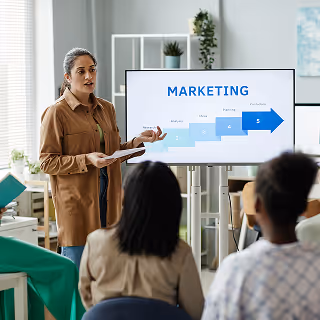Woman giving a marketing presentation to a small seated group, pointing to a digital screen that displays a five-step arrow diagram.