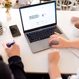 Two people work together at a white table, one typing on a laptop showing a website homepage, surrounded by coffee mugs and a phone.