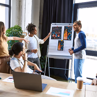 A group of young professionals gather around a flip chart as one member presents colorful data visualizations in a bright, modern meeting room.
