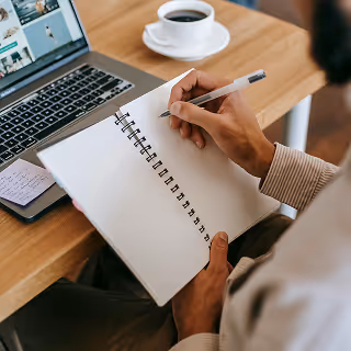 A person in a beige shirt writes in a spiral notebook while seated at a table with an open laptop, coffee cup, and notepad.