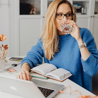 A blonde woman in glasses and a blue sweater drinks a glass of water while working at a white desk with a laptop, open notebook, and colorful office supplies.
