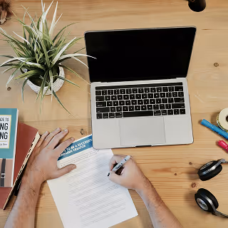 Overhead view of a person filling out a printed form on a wooden desk with a laptop, potted plant, headphones, and productivity books.