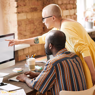 Two coworkers reviewing information on a desktop screen in a casual office setting, with one person pointing while the other navigates, suggesting collaborative decision-making.