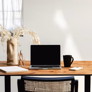 Minimalist workspace with a laptop, black mug, notepad, and decorative dried flowers on a wooden desk, evoking a calm and organized environment for remote work or planning.