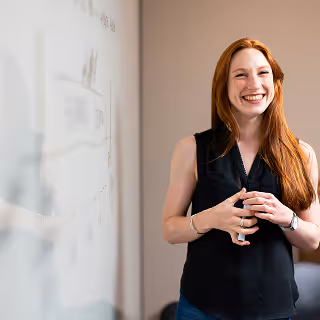 Confident woman standing in front of a whiteboard with diagrams, smiling and gesturing as if presenting or teaching, representing leadership and entrepreneurship.