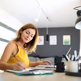 Focused woman working at a stylish desk, writing notes while referencing a laptop, illustrating planning and multitasking in a home office setting.