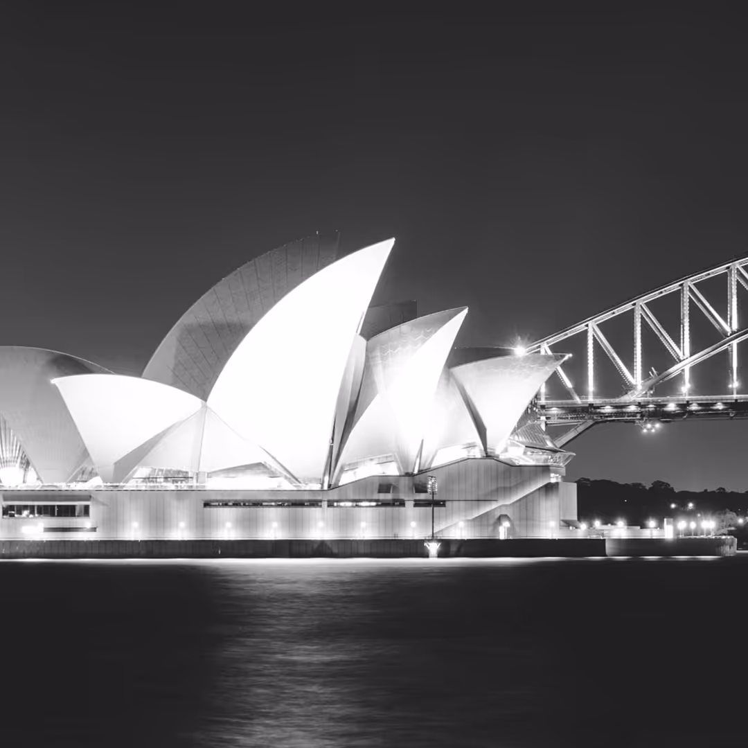 The Sydney Opera House illuminated against the night sky, with its iconic sail-shaped design and the Sydney Harbour Bridge in the background.