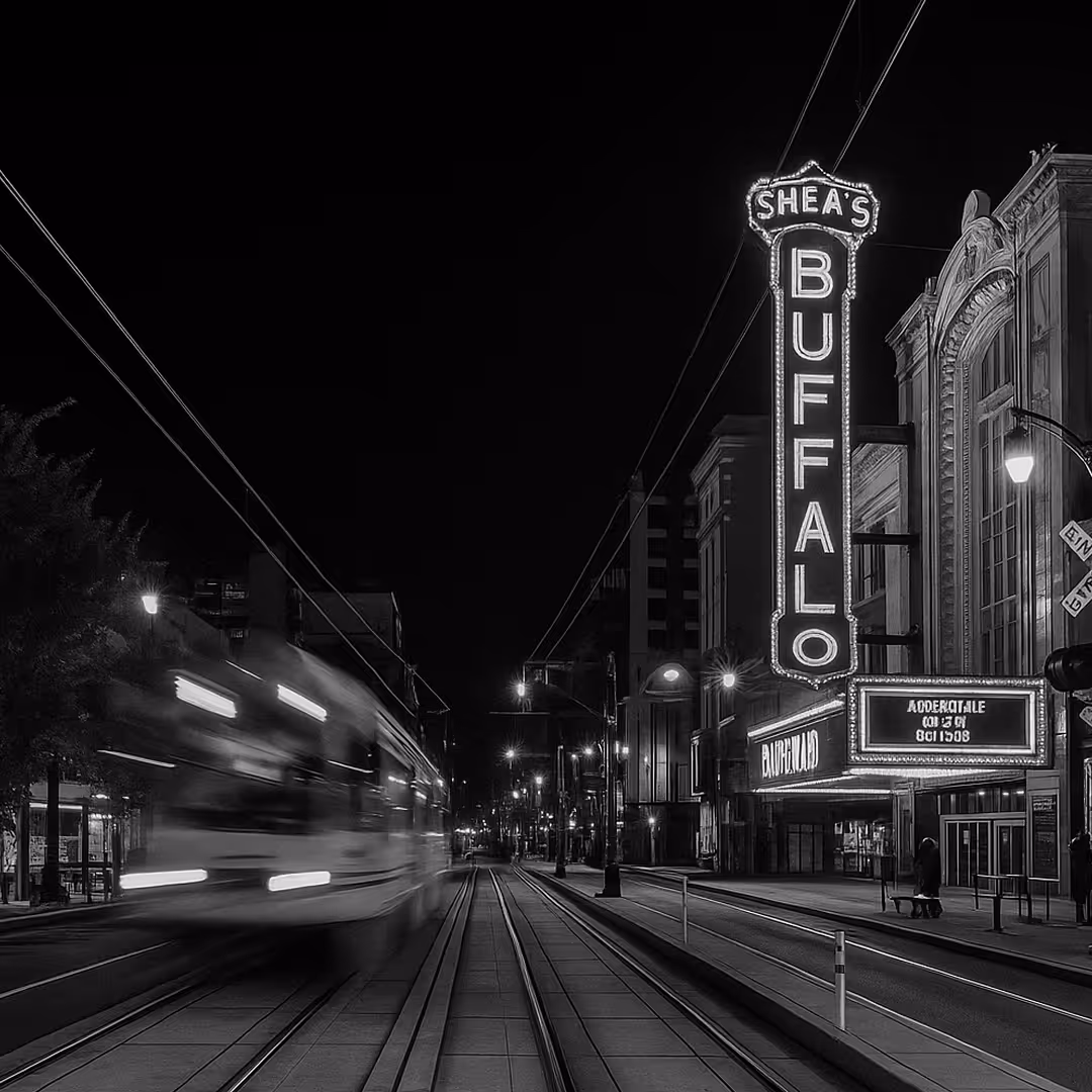 Nighttime street view showing the Shea’s Buffalo Theatre with its large illuminated vertical sign as a light rail passes by.