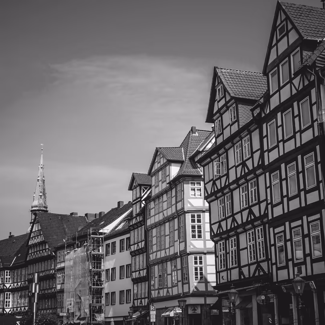 Black and white photo of traditional German half-timbered houses with steep roofs, tall windows, and a church steeple in the background under a partly cloudy sky.