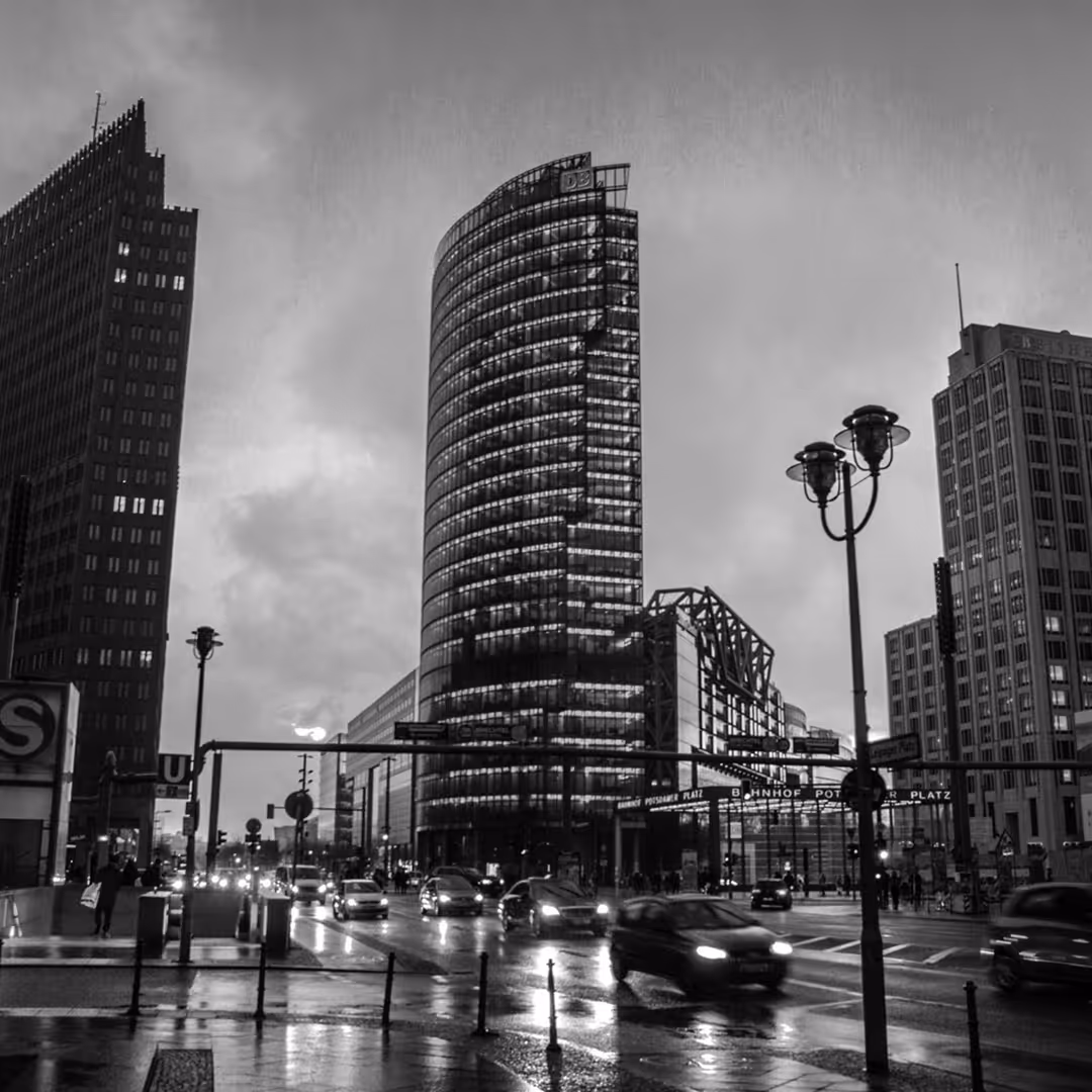 Black and white photo of a modern Berlin street scene with the curved DB Tower lit up, cars moving in the rain, and surrounding skyscrapers at dusk.