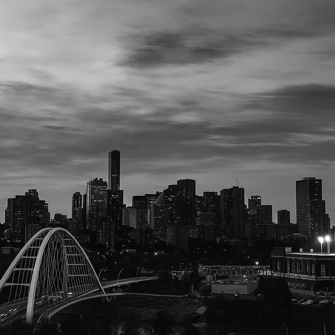A black-and-white photo of Edmonton’s skyline with high-rise buildings silhouetted against a dramatic evening sky, and a modern bridge leading into the city.