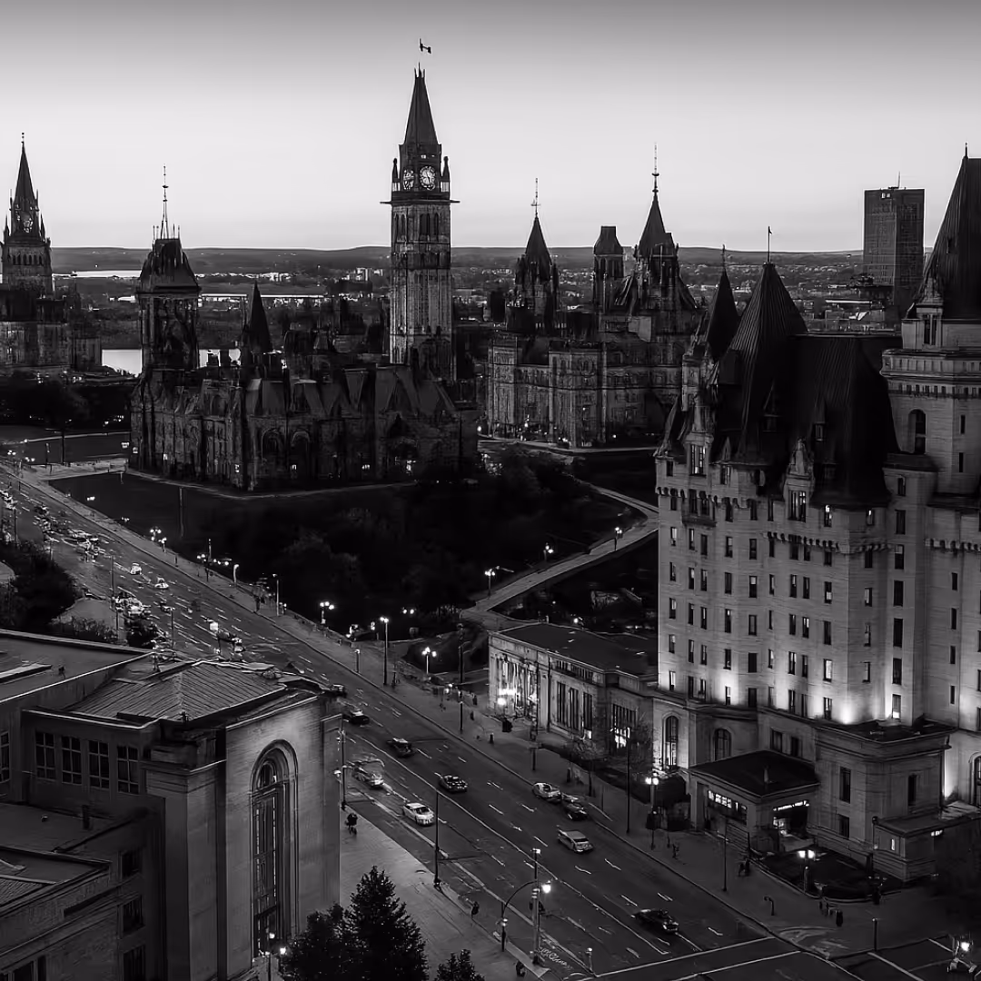 A dramatic black-and-white photo of Ottawa’s Parliament Hill, showing the historic Parliament buildings and Peace Tower illuminated as evening sets in.